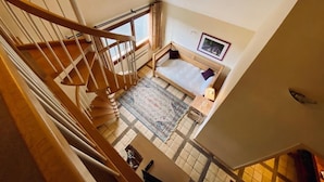 Une pièce aux hauts plafonds, avec un escalier en bois, un lit doté d’une tête de lit, une petite table et une fenêtre.
