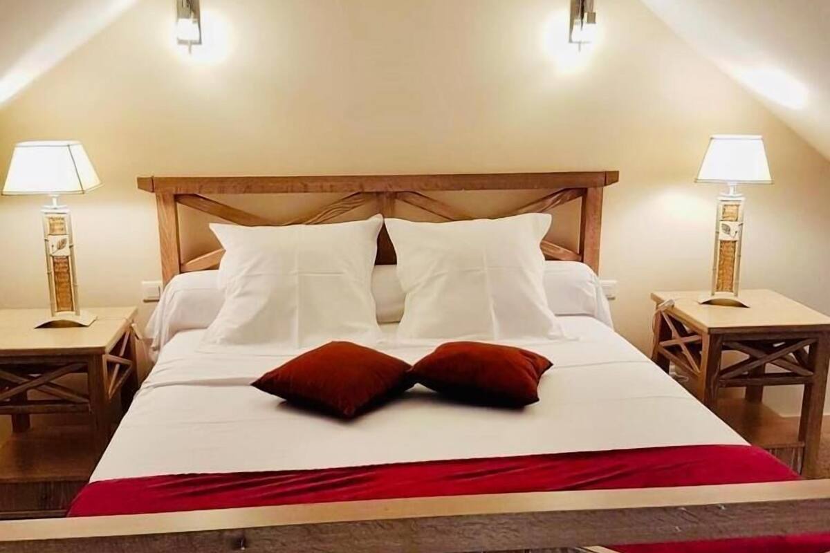 Un dormitorio con cabecera de madera, dos lámparas de noche, cama con sábanas blancas y dos almohadas rojas, y una manta roja.
