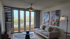 A modern living room with a sofa, a coffee table, and a floor lamp. There are large windows with blinds, a ceiling fan, and two paintings on the wall. - Your Cozy Combate beach retreat, 2 bed/2bath, full A/C, free laundry (Combate)