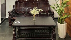 A room with a dark wooden bench, a small table with a vase of flowers, a wall-mounted fan, and a potted plant. - Shine Homestay Hue (Hue)