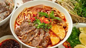 A bowl of beef noodle soup with sliced beef, tofu, and garnishes like green onions and chili peppers. - Shine Homestay Hue (Hue)