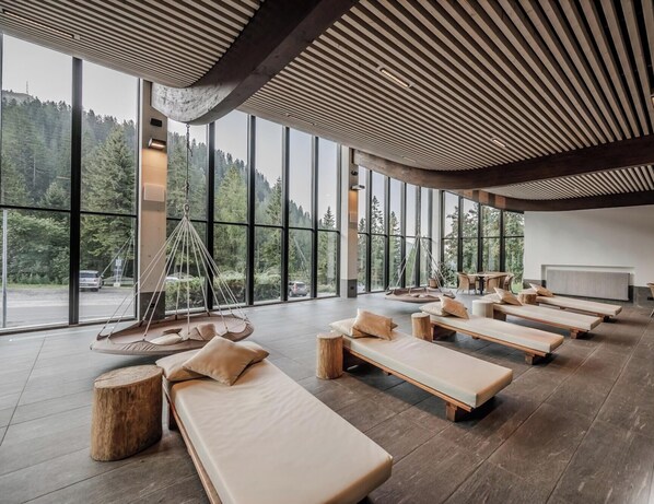 A modern spa room with large windows, wooden loungers, and a hanging chair. - HOTIDAY Apartment Collection - Campiglio Grostè (Pinzolo)