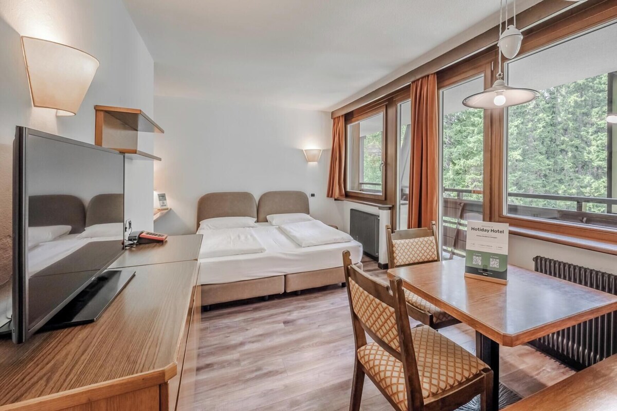 A hotel room with a bed, a desk, a chair, and a dining table. There are windows with curtains, a radiator, and a sign indicating the room type.