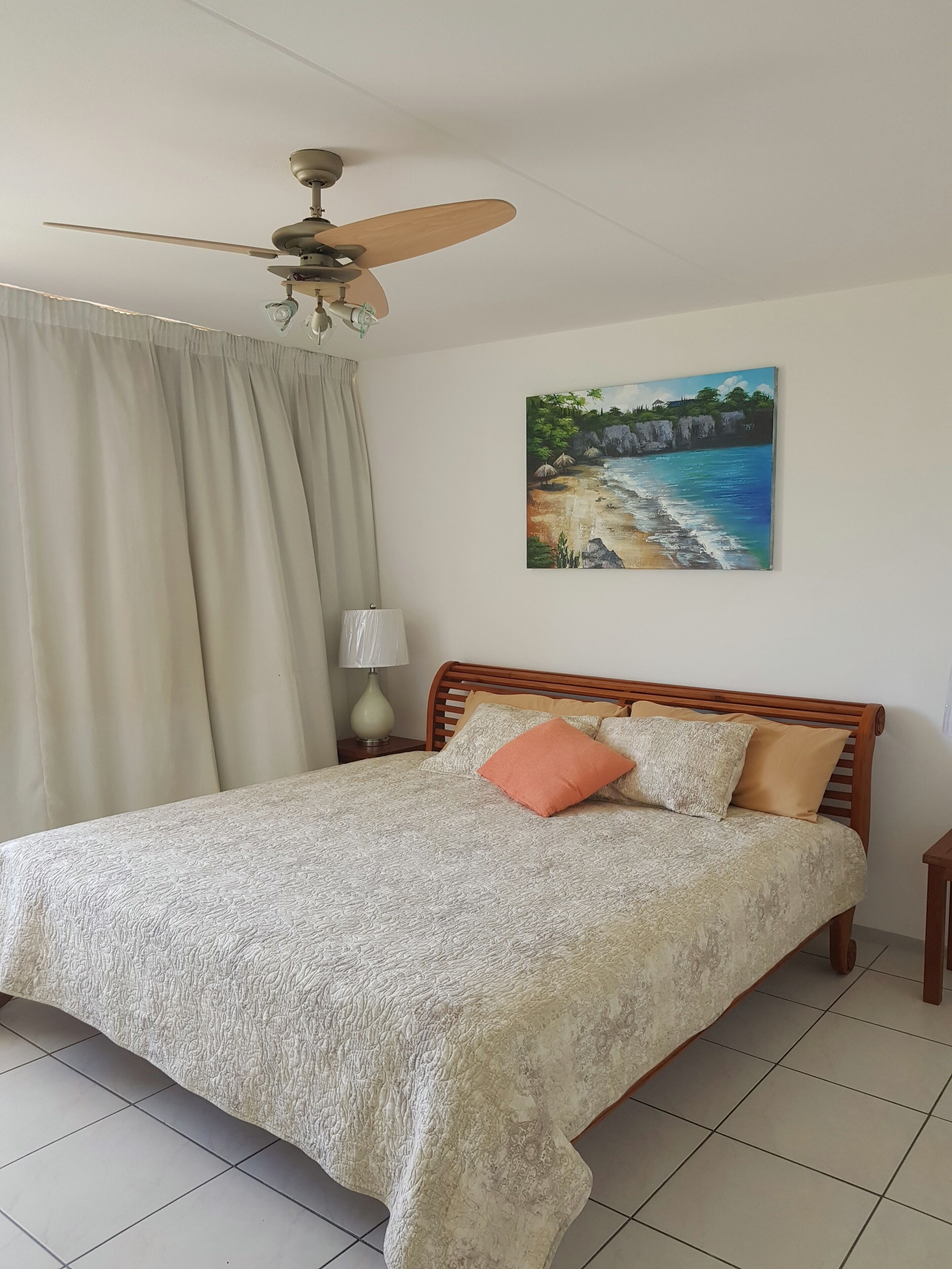 Una cama bien hecha con un edredón estampado, un ventilador de techo, un cuadro con una escena de playa y una lámpara en una mesita de noche.