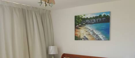 Una cama bien hecha con un edredón estampado, un ventilador de techo, un cuadro con una escena de playa y una lámpara en una mesita de noche.