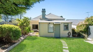 A small, single-story house with a sloped roof and a chimney. It has a well-maintained lawn and a small garden area with various plants and shrubs.