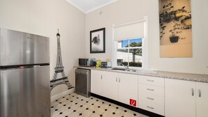 A kitchen with white cabinets, a stainless steel refrigerator, a microwave, and a sink. There's a framed picture of the Eiffel Tower on the wall, and a window with a view of trees and a house.