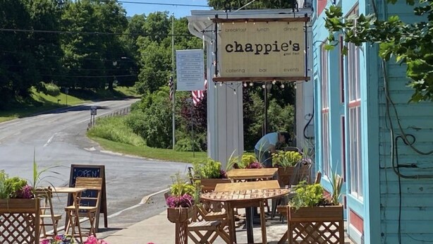 Un área de estar al aire libre con mesas y sillas de madera, un letrero que dice "chappie's" y vista a una calle.