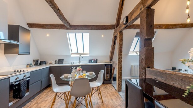 A modern kitchen with a dining area, featuring exposed wooden beams, a large window, and a built-in oven. - 5C Charming Town Centre Apartment With Roof Top Views - Pass the Keys (Shrewsbury)