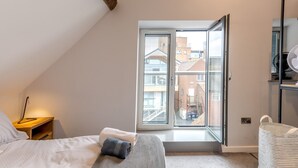 A bedroom with a bed, a bedside table with a lamp, a window with a view of buildings, and a fan on a shelf. - 5C Charming Town Centre Apartment With Roof Top Views - Pass the Keys (Shrewsbury)