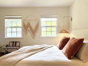 Une chambre avec un lit, deux fenêtres, une décoration murale en macramé, une lampe sur pied et une chaise.