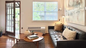 A living room with a leather sofa, a coffee table with books, a large window with blinds, and a wall-mounted artwork. - the hideaway: w/pool, hot tub, relaxing courtyard (Houston)