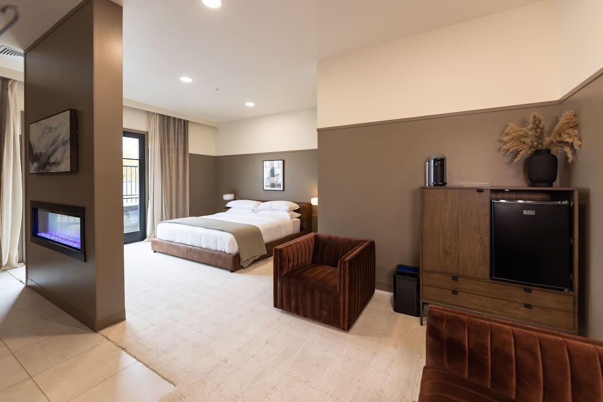 A modern hotel room with a bed, a seating area, a TV, and a minibar.