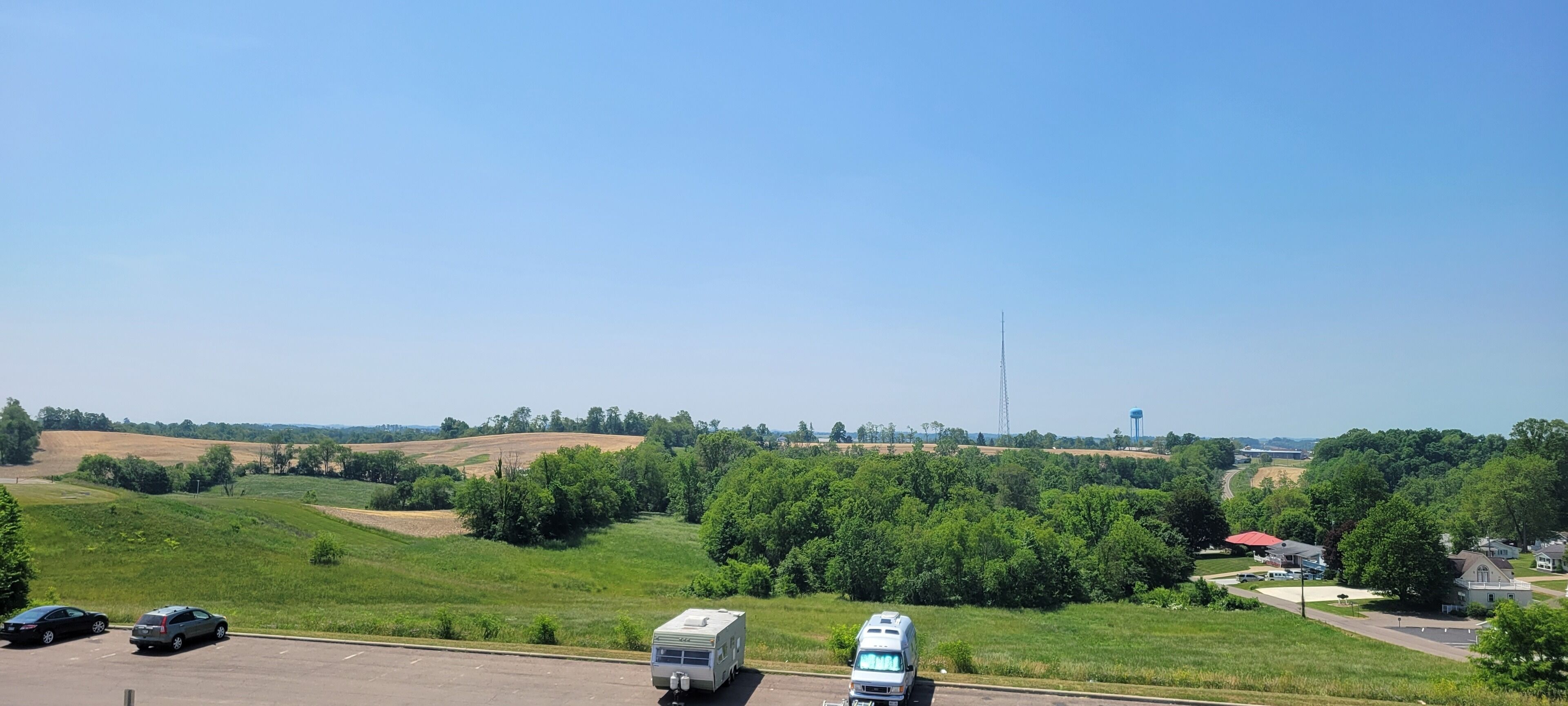 Ein Parkplatz mit Wohnmobilen und Autos, umgeben von grünen Feldern und Bäumen.