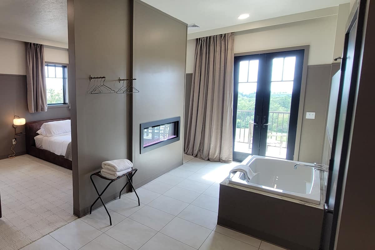 A modern hotel room with a large bed, a sitting area with a table, a bathroom with a bathtub, and a view of greenery outside.