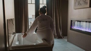 A person in a white robe standing by a bathtub in a room with a large window and a wall-mounted aquarium. - The Atlee Suites (Berlin)