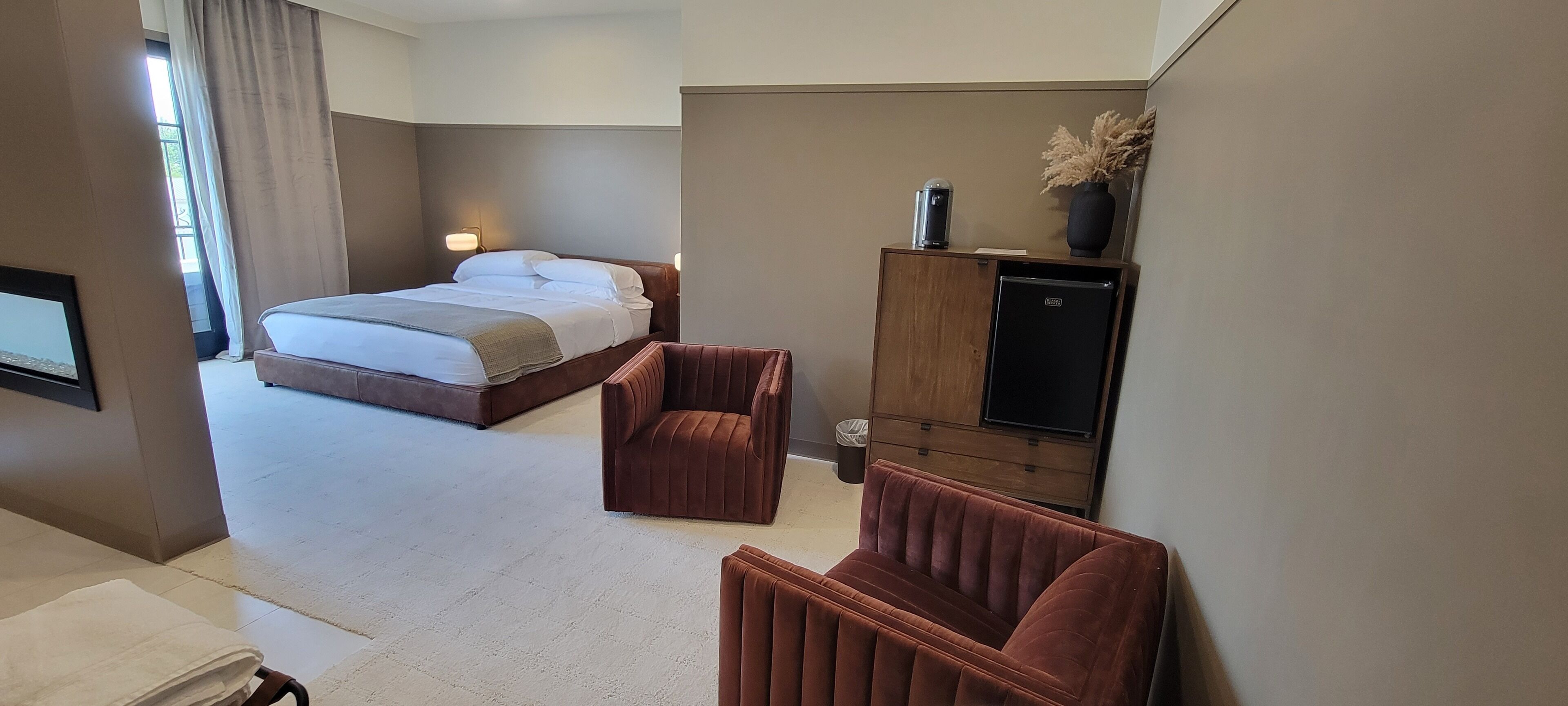 A hotel room with a bed, two armchairs, a TV, a minibar, and a small cabinet.