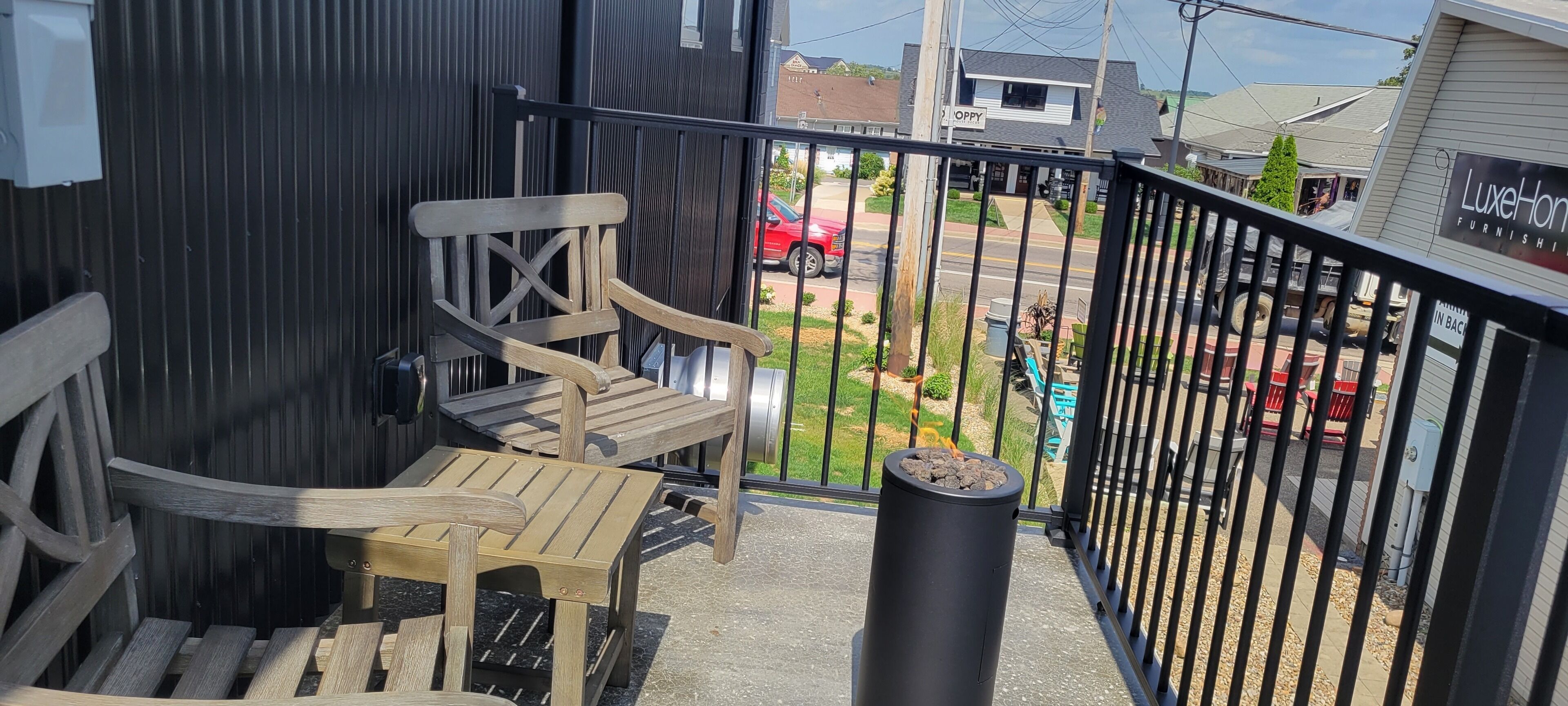 A balcony with wooden chairs and a table, a black trash can, and a view of a street with a red car and a building labeled 'LUXE Home Furnishings'.