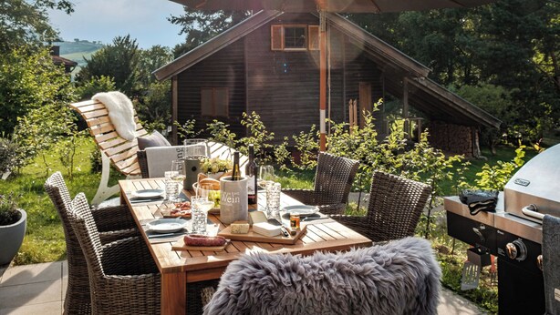 An outdoor dining area with a wooden table set for a meal, surrounded by wicker chairs, a grill, and a large umbrella providing shade. - Ferienhaus Sonnenweg 32 - Sauna, Garten und Seeblick Inklusive (Diemelsee)