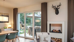 A modern living room with a fireplace, a dining area with a table and chairs, and a large sliding glass door leading to an outdoor patio. - Ferienhaus Sonnenweg 32 - Sauna, Garten und Seeblick Inklusive (Diemelsee)