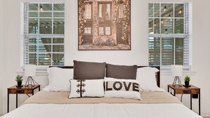 A bedroom with a large bed, two nightstands, a wall-mounted painting, and two windows with blinds. - Forest Retreat - BBQ - Game Room (Shreveport)