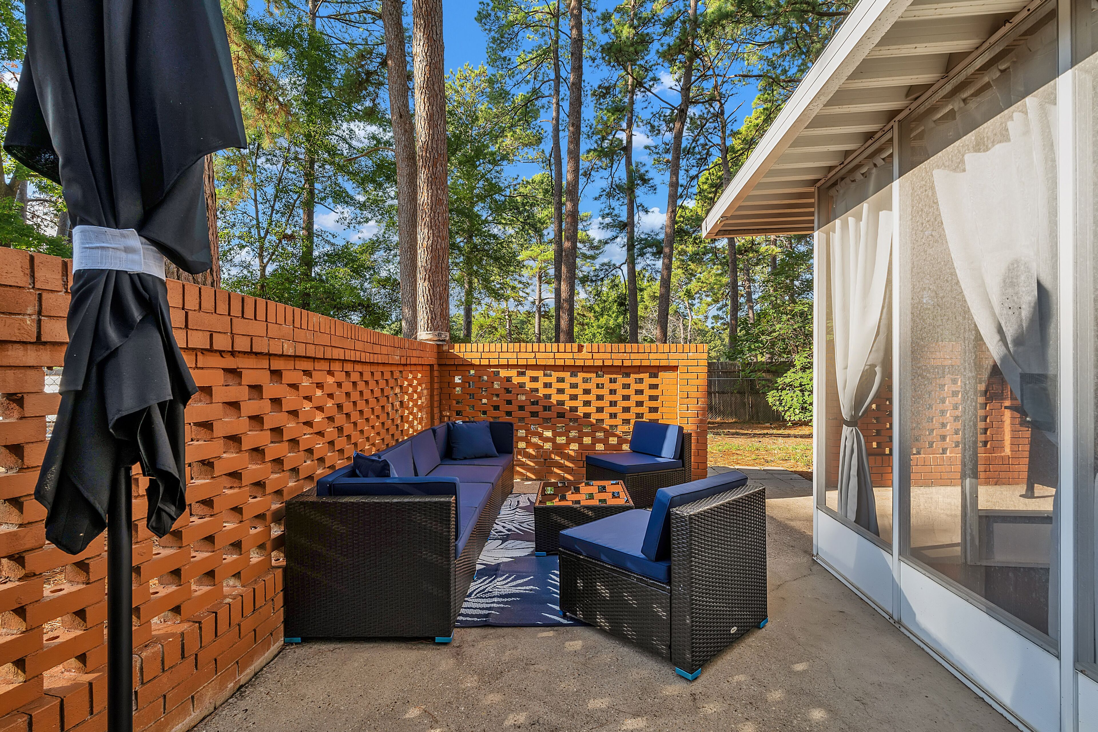Un espacio de patio al aire libre con mobiliario de mimbre, pared de ladrillo y puerta de cristal que da a un área techada.