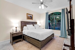 Une chambre avec un lit en bois, deux tables de chevet avec des lampes, un ventilateur de plafond, une grande fenêtre avec des rideaux et un tableau accroché au mur.