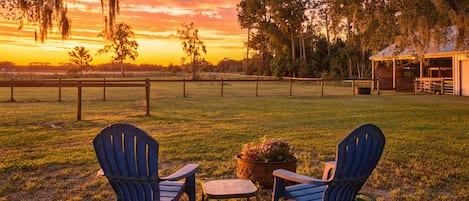 Une vue au coucher du soleil, avec deux chaises et une table dans un champ herbeux, entouré d’arbres et d’une clôture.