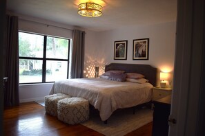 Ein Schlafzimmer mit großem Fenster, einem Bett mit Kopfteil, einem Nachttisch mit Lampe und einem Wanddekor.