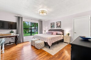 A bedroom with a bed, bedside tables, a TV, a window with curtains, and a view of greenery outside.