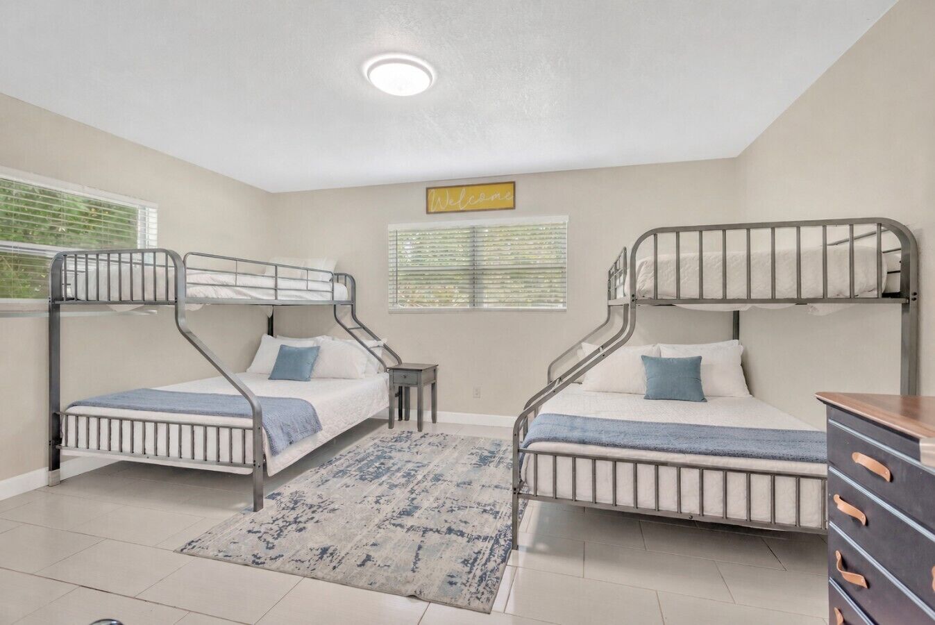 A room with bunk beds, a small table, a dresser, and a window with blinds.