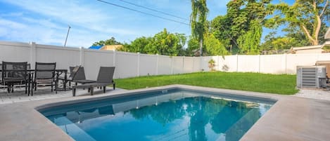 A rectangular swimming pool with a white fence and a patio area with chairs and a table.