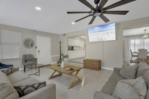 A modern living room with a ceiling fan, a television, and a view of the beach through the window.