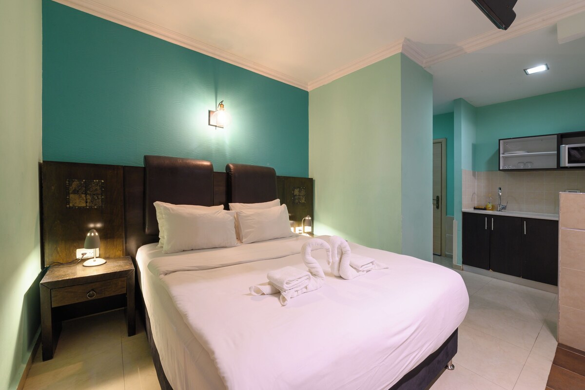 A hotel room with a large bed, a nightstand with a lamp, a wall-mounted light, and a kitchenette area.