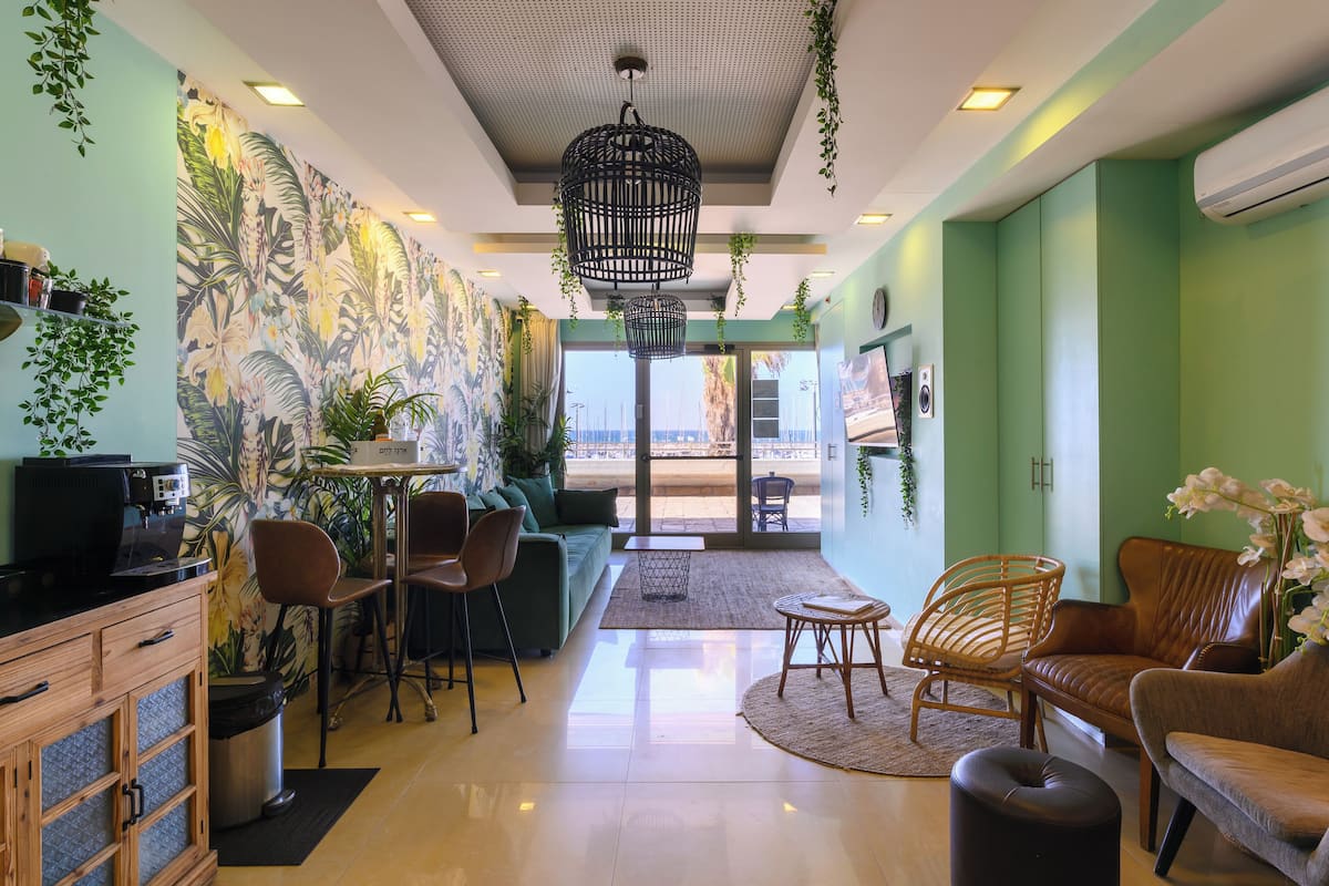 A modern interior with a green wall, tropical wallpaper, a coffee machine, seating area, and a view through a glass door.