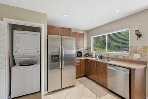 Una cocina con un refrigerador de acero inoxidable, horno empotrado, lavavajillas, armarios de madera, encimera de granito y una ventana con persianas.