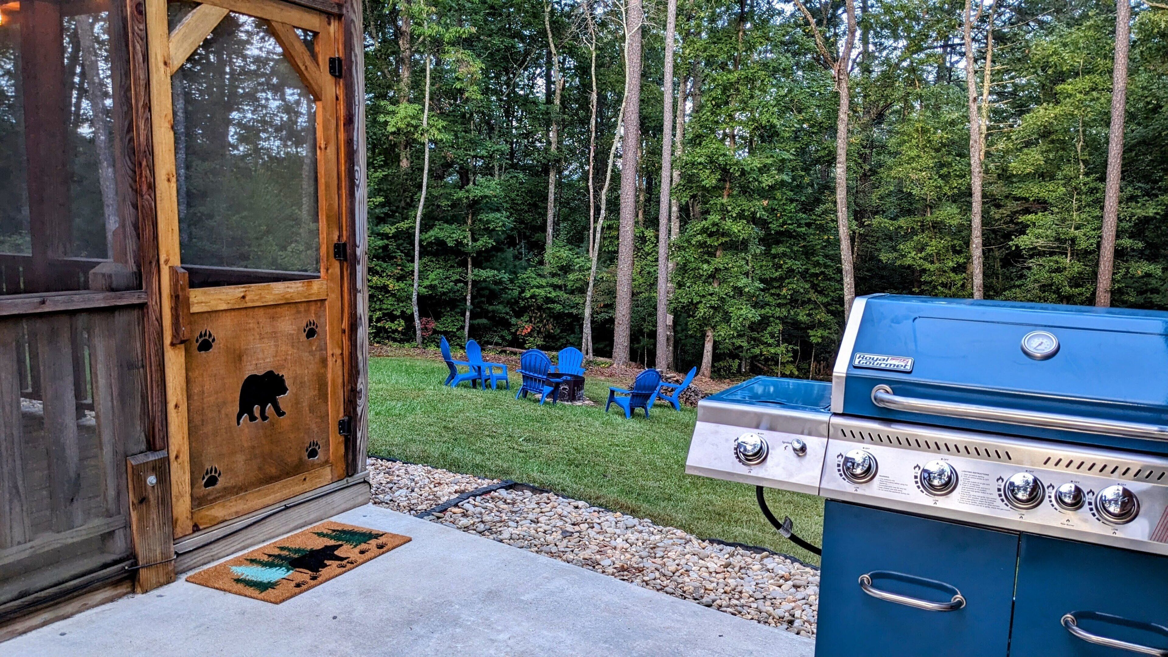 A wooden door with bear paw prints, a blue grill, and blue Adirondack chairs in a grassy yard with trees.