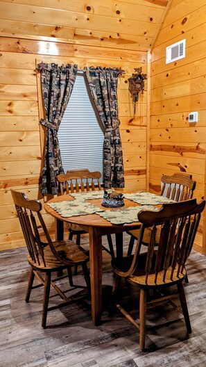 A wooden cabin interior with a round dining table, six wooden chairs, patterned curtains, and a decorative clock.