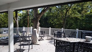 Una terraza techada al aire libre con barandillas blancas, sillas y mesas de metal negro, rodeada de árboles.