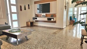 A modern living room with a large flat-screen TV mounted on the wall, a grey sofa, a coffee table with magazines, and a speckled floor. - San Juan Charm • ForestEdge Retreat • Gated Privacy Near Beach, Airport & Old SJ (San Juan)