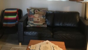A living room with a black leather sofa, a colorful blanket, a patterned pillow, a wooden table with decorative items, and a window with white curtains. - Lake front bachelor 
Close to all amenities casino Rama 
Ski hills  (Orillia)