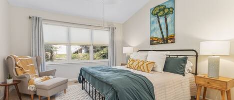 A bedroom with a bed, a chair, a nightstand, a lamp, a painting, and a window with curtains.