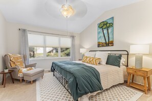 Une chambre à coucher comprenant un lit, une chaise, une table de chevet, une lampe, un tableau et une fenêtre avec des rideaux.