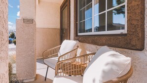 A balcony with wicker chairs and a white cushion, a window with a view of the sky, and a textured wall.