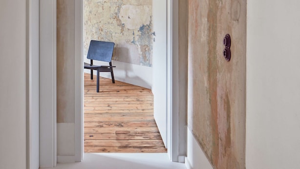 Un couloir étroit, doté d’un plancher en bois, mène à une pièce dont le mur est défraîchi et qui contient une chaise bleue.