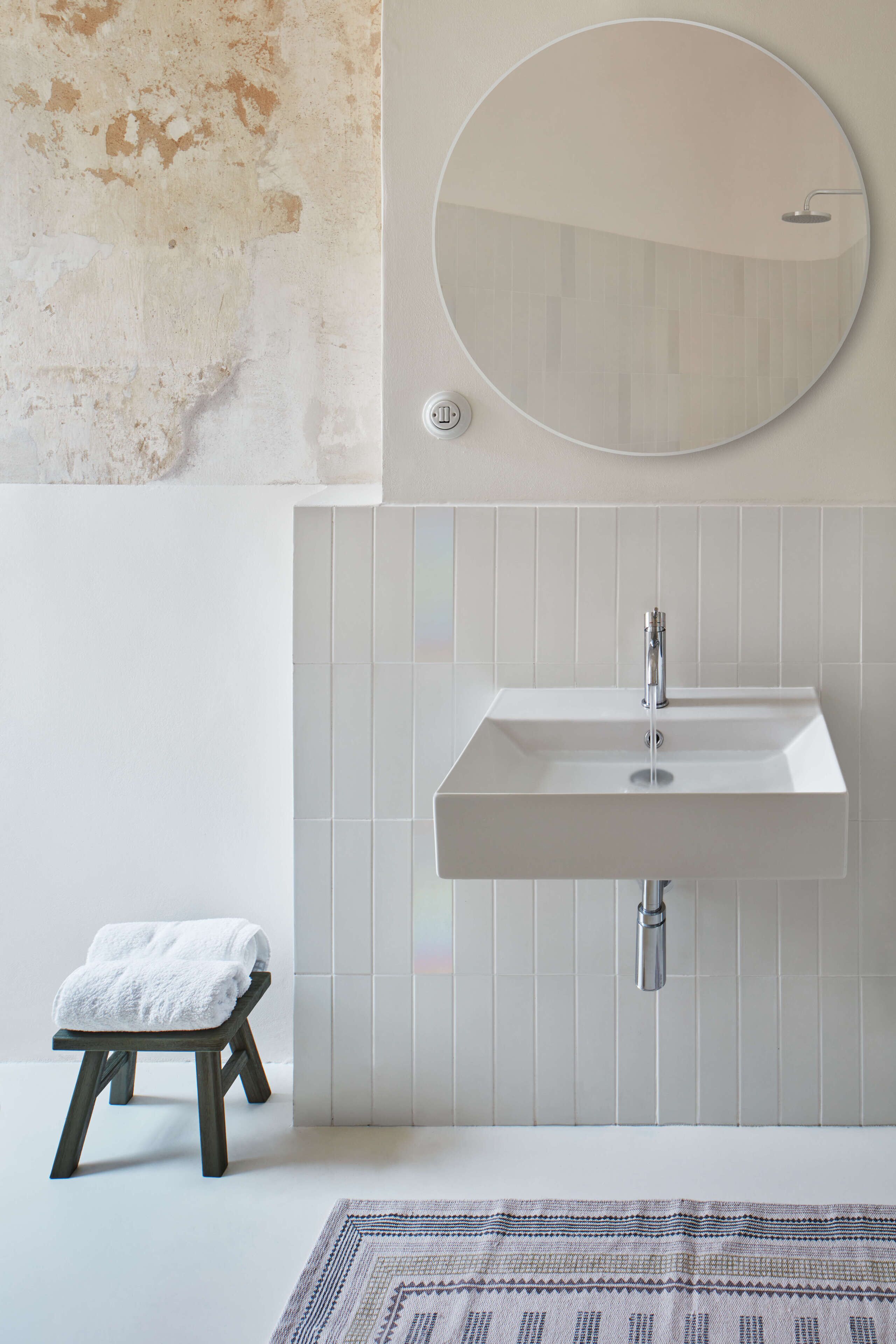 A bathroom with a rectangular sink, a round mirror, and a small stool with a towel.