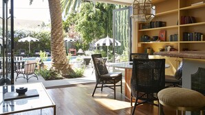 A sunlit room with a view of an outdoor pool area, featuring a wooden table, chairs, and a bookshelf.