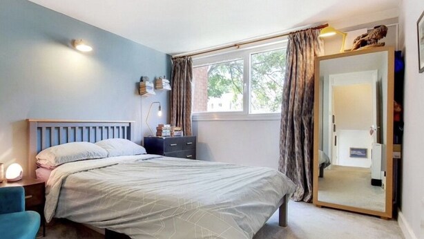 A bedroom with a bed, bedside table, lamp, and a window with curtains. - Bakersfield (London)