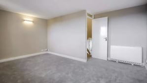 A room with a grey carpet, white walls, a white radiator, and a door leading to another room. - Bakersfield (London)