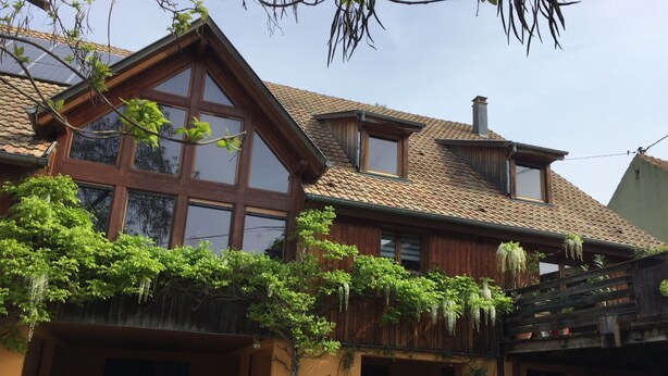 A two-story wooden house with large windows, a tiled roof, and a covered porch area.
